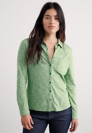 Mladá žena s dlouhými tmavými vlasy, oblečená ve světle zelené košili na knoflíky a tmavých kalhotách, stojící před světlým jednobarevným pozadím.