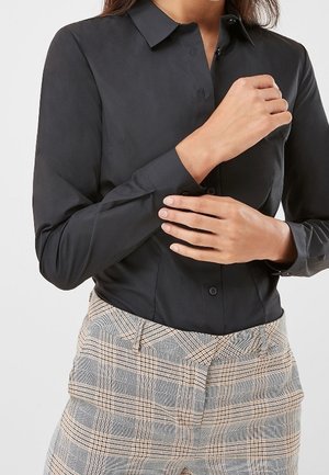 Personne portant une chemise noire ajustée à manches longues boutonnée et un pantalon à carreaux taille haute ajustant le poignet de la chemise.