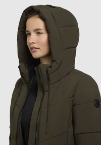 Zateplená bunda v olivově zelené barvě s vysokým límcem, velkou kapucí, černým zipem a nastavitelnými šňůrkami. Má strukturovaný povrch a kulatou nášivku.