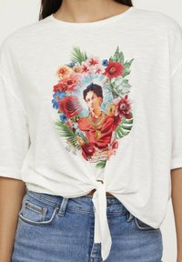 T-shirt blanc à l'avant noué, avec un imprimé floral coloré et un design central représentant une femme aux cheveux foncés et aux accessoires en or.