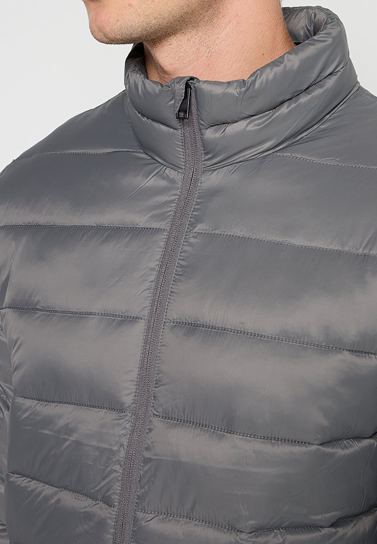 Chaqueta de plumón gris con cuello alto, cierre frontal con cremallera y textura acolchada horizontal. Fabricada con un tejido ligero y brillante.