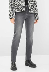 Persona che indossa jeans slim fit grigi, stivali neri voluminosi e una giacca con stampa leopardata bianca e nera, in piedi su uno sfondo bianco.