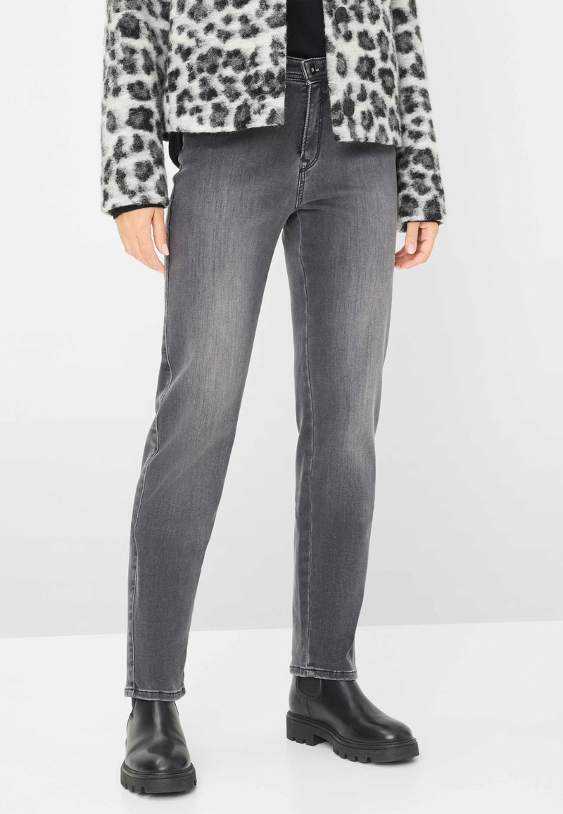 Persona che indossa jeans slim fit grigi, stivali neri voluminosi e una giacca con stampa leopardata bianca e nera, in piedi su uno sfondo bianco.