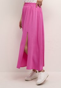 Longue jupe rose avec une fente, taille élastique et tissu léger, associée à des baskets blanches sur un fond clair.