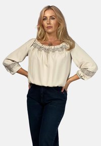 Kremowa bluzka z haftowanym koronkowym wykończeniem przy dekolcie i rękawach. Posiada rękawy o długości 3/4 oraz luźny krój, zestawiona z ciemnymi dżinsami.