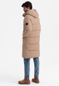Abrigo tipo pluma beige con capucha, que presenta un diseño acolchado horizontal, puños elásticos y un parche negro en la manga. Llevado sobre jeans azules y botas marrones.