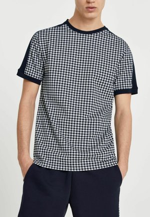 Mann trägt ein kurzärmeliges Hemd mit schwarz-weißem Hahnentritt-Muster, einfarbige schwarze Ärmel und schwarze Hose, Hände in den Taschen.