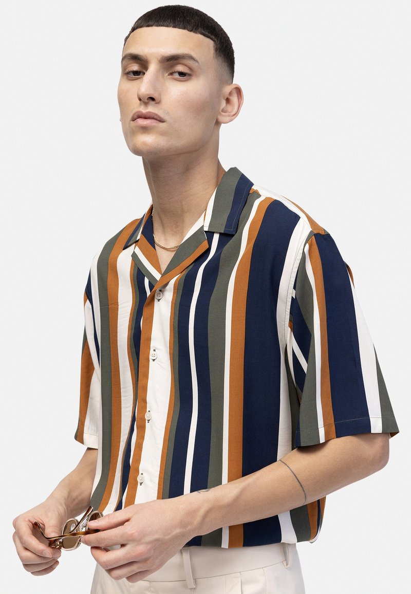 Ung man som håller solglasögon och bär en kortärmad skjorta med knappar och vertikala ränder i blått, orange, vitt och grönt.