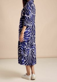 Blå och vit mönstrad klänning med lös passform, långa ärmar och flerlagersdesign, i kombination med vita sneakers på en neutral bakgrund.