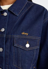 Tamno plava traper jakna s daščicama u srebrnoj boji, zlatnim vezom "obey" i dva džepa na prsima sa kontrastnim šavovima.