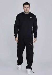 Črni pulover z okroglim izrezom z logotipom na prsih, kombiniran s črnimi cargos hlačami z žepi in zlatim poudarkom. Model nosi bele superge.