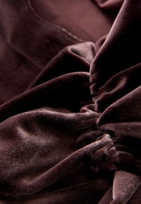 Tumši brūna samta audums ar mīkstu tekstūru, ar savākiem posmiem, kas veido plūstošu izskatu un smalku spīdumu.