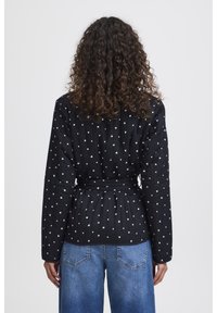 Veste matelassée noire avec des motifs de cœurs blancs, cintrée à la taille, comportant des manches longues et un col classique. Associée à un jean bleu.