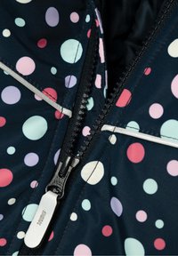 Gros plan sur une veste bleu marine partiellement zippée, ornée de pois multicolores et d'une languette réfléchissante sur le zip.