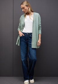 Hellgrüner langer Cardigan, weißes Shirt, dunkelblaue Jeans und weiße Turnschuhe. Der Cardigan hat gerippte Bündchen; die Jeans sind ausgestellt. Das Modell steht vor einem grauen Hintergrund.