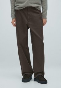 Brune brede bukser lavet af glat stof, med knaplukning og sidelommer, parret med mørke slip-on sko.