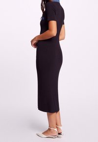 Kvinna klädd i en figurnära svart midiklänning med korta ärmar och blå detaljer vid kragen, som hon har parat med vita lågklackade ankelremsskor.