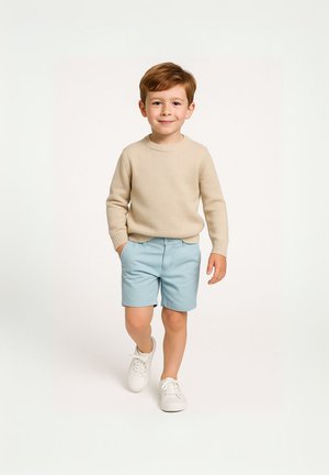 Maglione lavorato a maglia beige, pantaloni blu, sneakers bianche, bambino in piedi con i capelli corti castani su uno sfondo chiaro. Outfit semplice e informale.