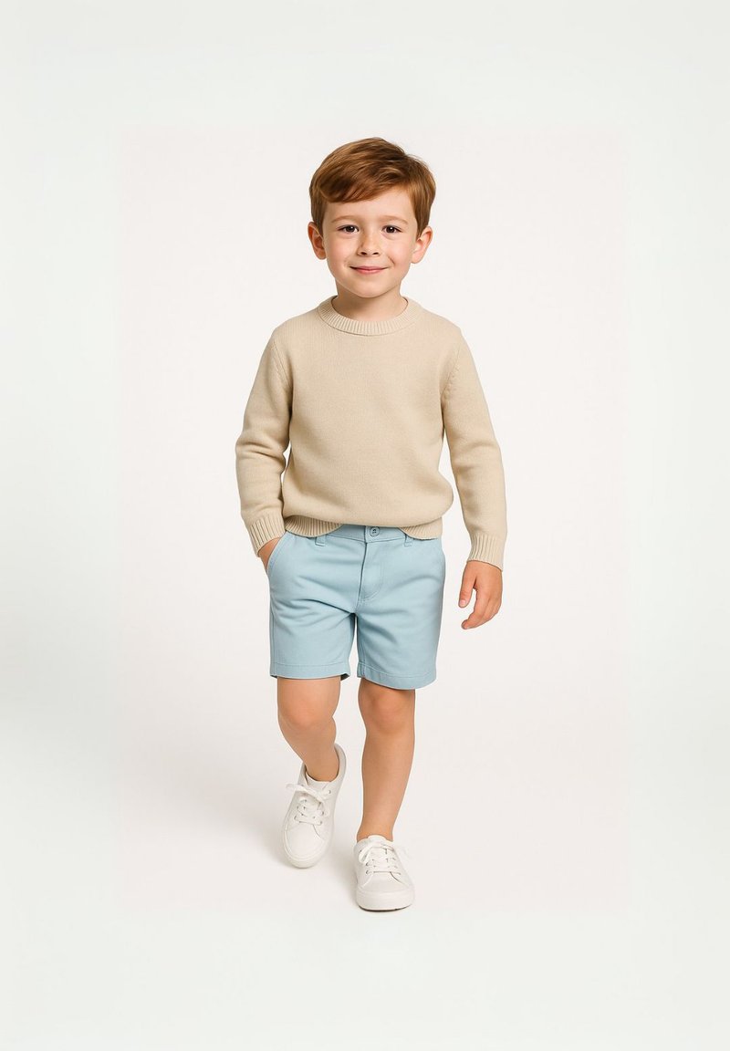 Bežový pletený sveter, modré kraťasy, biele tenisky, stojaci dieťa s krátkymi hnedými vlasmi na svetlom pozadí. Jednoduchý, neformálny outfit.