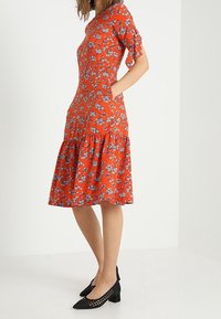 Röd blommig klänning med korta ärmar, påsydd midja, utställd nederkant och sidofickor. Svarta, perforerade klackskor med blockklack kompletterar stilen.