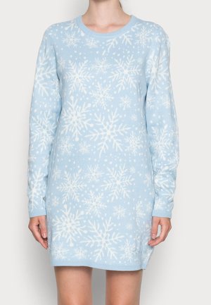 Woman wearing light blue long-sleeve sweater dress with white snowflake and dotted patterns, standing against a plain background.