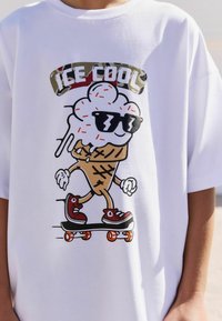 Witte T-shirt met een cartoon-ijsje dat een skateboarder is, met een zonnebril op, en de tekst "ICE COOL" in camouflageprint erboven.