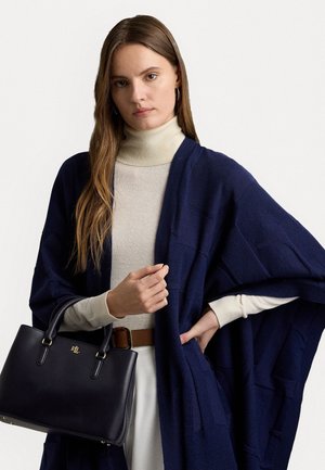 Jeune femme aux longs cheveux bruns porte un col roulé beige et un châle bleu marine, tenant un sac à main en cuir noir avec des détails dorés.