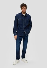 Donkerdenim overhemd met knoopsluitingen, twee borstzakken en een rechte pasvorm, gecombineerd met donkere jeans en witte sneakers.
