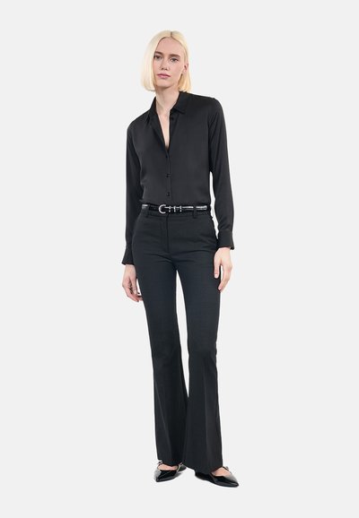 Blusa di seta nera con bottoni frontali, abbinata a pantaloni neri aderenti in tessuto strutturato, impreziositi da una cintura e ballerine nere.