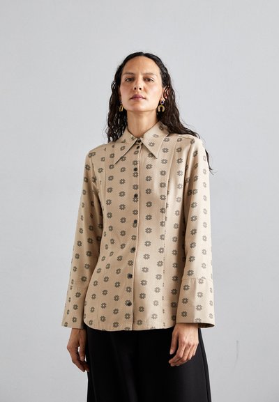 Beige skjorte med blomstermønster, der har spidse kraver, lange ærmer og sorte knapper langs forsiden. Glat stofstruktur.