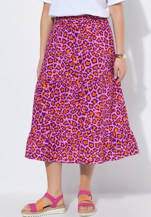Frau trägt einen Midi-Rock mit Leopardenmuster in Pink, Lila und Orange, dazu orange-pinke Sandalen und ein weißes Oberteil, steht vor einem weißen Hintergrund.