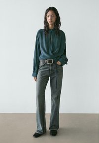 Tyrkysově modrá saténová halenka s výstřihem do tvaru klíčové dírky, zavazovaná v pase, v kombinaci s vysokým pasem šedých zvonových džínů. Černé lodičky s uzavřenou špičkou.