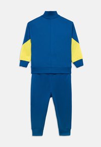 Μπλε φόρμα με ψηλό γιακά, κίτρινα διακοσμητικά στις μανσέτες και στενόξυλα παντελόνια με ρεβέρ, κατασκευασμένη από απαλό, ελαστικό υλικό.