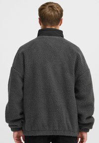 Grå fleecejakke med høj krave, der har en tekstureret overflade og drop shoulder-ærmer. Designet inkluderer en elastisk kant og manchetter.