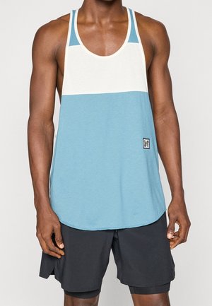 Gespiert persoon met een mouwloos blauw-wit tanktop en zwarte sportshort, staand tegen een effen lichte achtergrond.