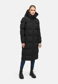 Black puffer coat with a long silhouette, padded design, adjustable hood, and zipper closure, paired with blue jeans and black boots.