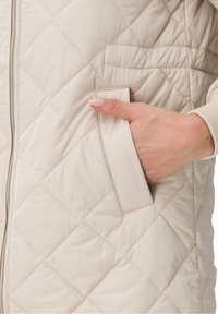 Gilet trapuntato beige con chiusura a zip e tasca laterale. Il tessuto ha una texture liscia con un motivo a rombi e una finitura morbida.