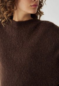 Rjav pleten pulover z visokim izrezom, mehko teksturo in širokimi rokavi, ki poudarjajo ohlapen, udoben kroj.