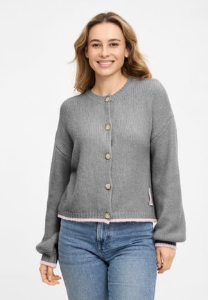 Frau lächelt, trägt eine graue, geknöpfte Strickjacke mit pinkfarbenem Saum und blaue Jeans vor weißem Hintergrund.