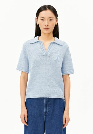 Jeune femme aux cheveux noirs et lisses portant un top en maille bleu clair à manches courtes avec col et un pantalon bleu à jambes larges.