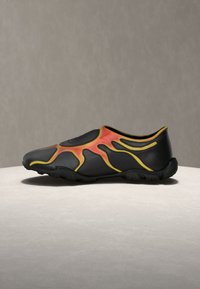 Μαύρο παπούτσι χωρίς κορδόνια με κόκκινες και κίτρινες φλόγες, ευέλικτη σόλα, τοποθετημένο σε στρογγυλή, ανοιχτόχρωμη επιφάνεια πάνω σε μονόχρωμο φόντο.