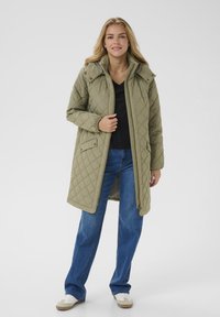 Kvinde iført en knælængde olivengrøn quilted frakke, sort skjorte, blå jeans og hvide sneakers, der står mod en ensfarvet lys baggrund.
