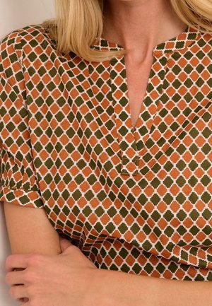 Femme portant un haut à motifs avec des formes géométriques orange et vertes et un col en V court, les bras croisés à la taille.