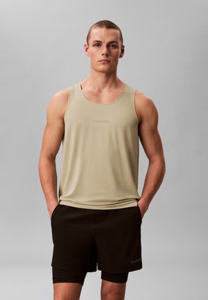 Joven de pie con las manos en los bolsillos, vestido con una camiseta sin mangas beige y pantalones cortos negros en capas, contra un fondo gris liso.