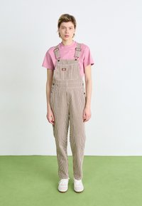 Trabajos de rayas en tela beige y blanca, con tirantes ajustables, un bolsillo en el frente y un corte relajado, combinados con una camiseta rosa.