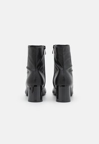 Une paire de bottines en cuir noir avec des talons carrés, vues de l'arrière sur un fond blanc.