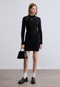 Μαύρο ριμπ φόρεμα με μακριά μανίκια και ψηλό γιακά, με λευκές ραφές. Το μοντέλο κρατά μια μικρή μαύρη τσάντα και φορά μαύρα μοκασίνια με λευκές κάλτσες.