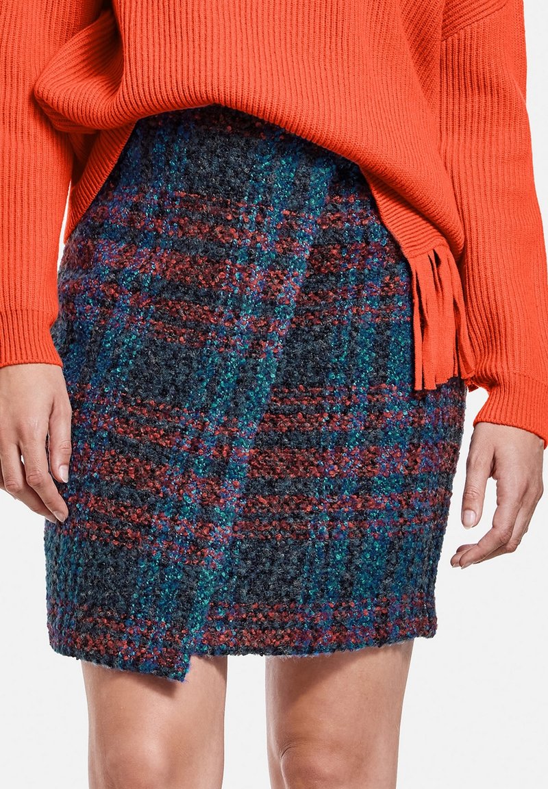 Femme portant une jupe en laine à carreaux texturée dans des tons bleu et rouille avec un pull côtelé orange avec des fentes latérales et des détails à franges.