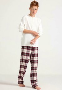 Haut blanc à manches longues avec un col henley, associé à un pantalon de pyjama beige et bordeaux à motifs à carreaux. Léger, coupe décontractée. Pieds nus.