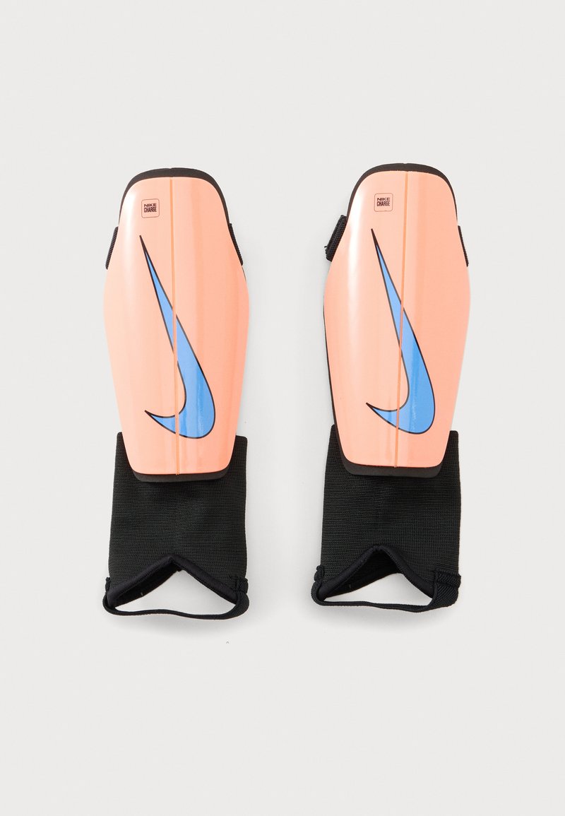 Ζευγάρι θωρακίσεις κνήμης Nike σε ροδακινί χρώμα με μπλε λογότυπο swoosh και προσαρτημένες μαύρες προστατευτικές μανσέτες για τον αστράγαλο, εμφανισμένες σε λευκό φόντο.